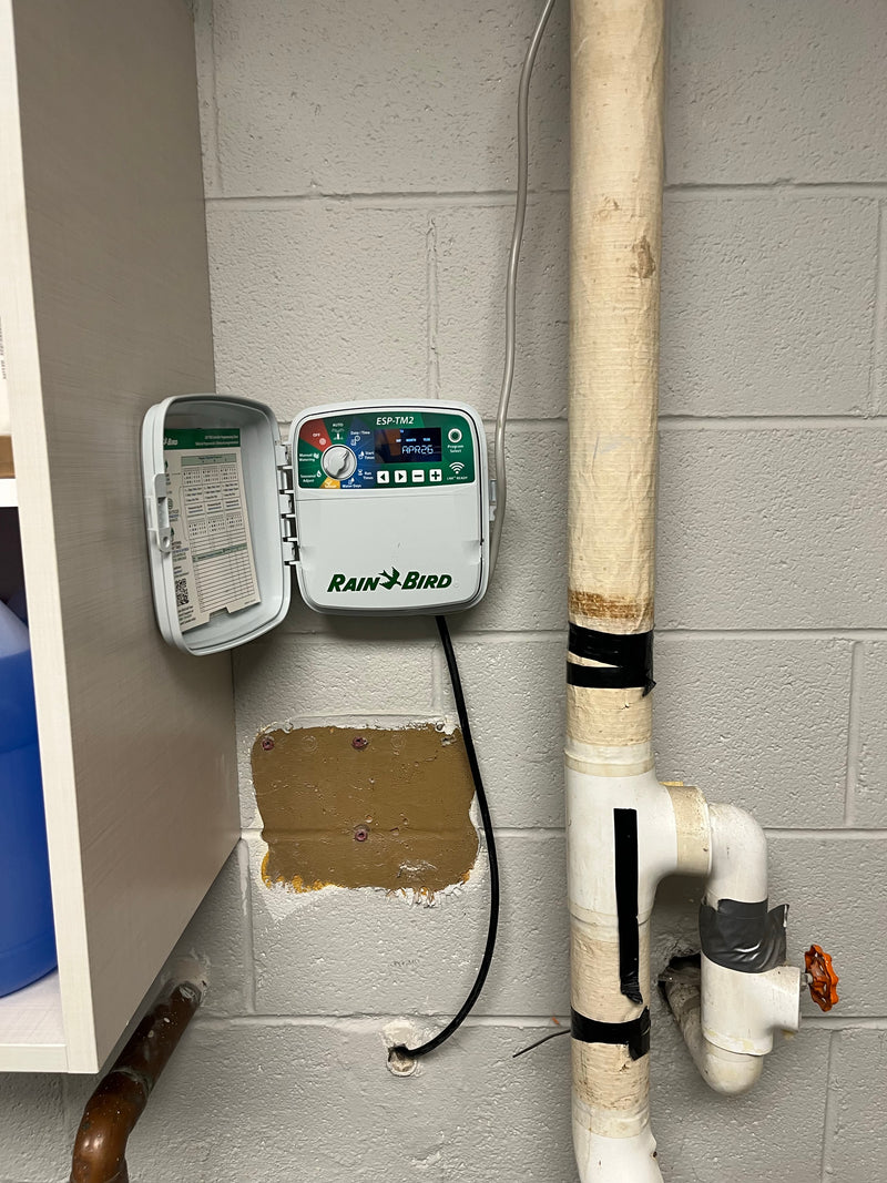 A Rain Bird sprinkler timer mounted on a gray cinderblock wall next to a vertical PVC pipe, with the timer's door open to show the settings.