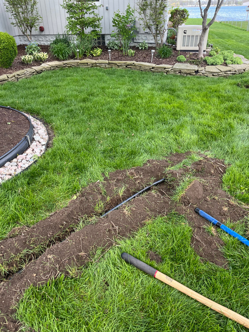 A backyard landscape scene with a freshly dug trench in the grass, a blue-handled tool, and a long-handled tool resting on the lawn. The background shows a landscaped yard with stone edging, plants, and a white fence.