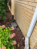 A close-up outdoor shot of a beige brick wall corner with a white downspout, mulch ground cover, a small black metal fixture, and pink hydrangea flowers in the foreground.