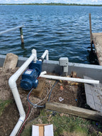 A blue pump motor connected to white PVC piping, installed outdoors on a wooden dock next to a body of water with a wooden pier visible.