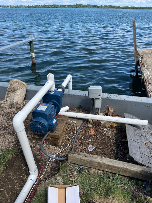 A blue pump motor connected to white PVC piping, installed outdoors on a wooden dock next to a body of water with a wooden pier visible.