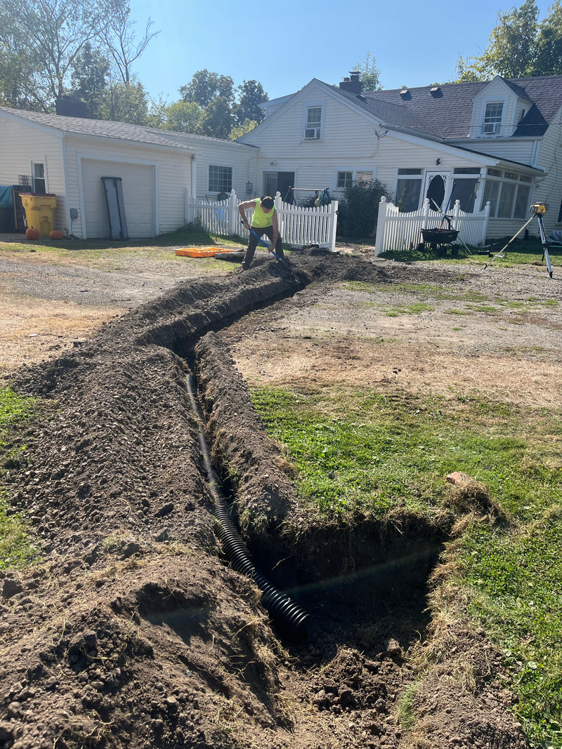 A trench for a drainage or sewer line is being dug in a residential backyard, with a worker standing at the top of the trench near a house and white picket fence.