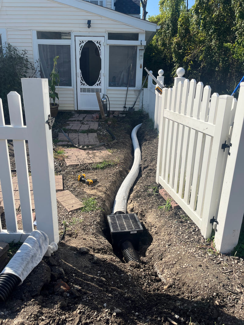 White PVC drain pipe installed in a trench next to a white picket fence in front of a house.