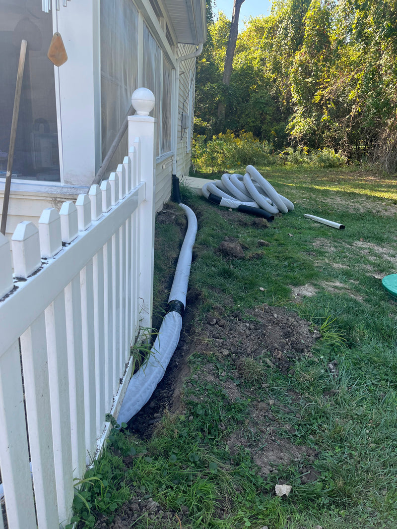 White PVC drainage pipe installed along the foundation trench of a house next to a white picket fence, with additional pipes stacked on the grassy yard.